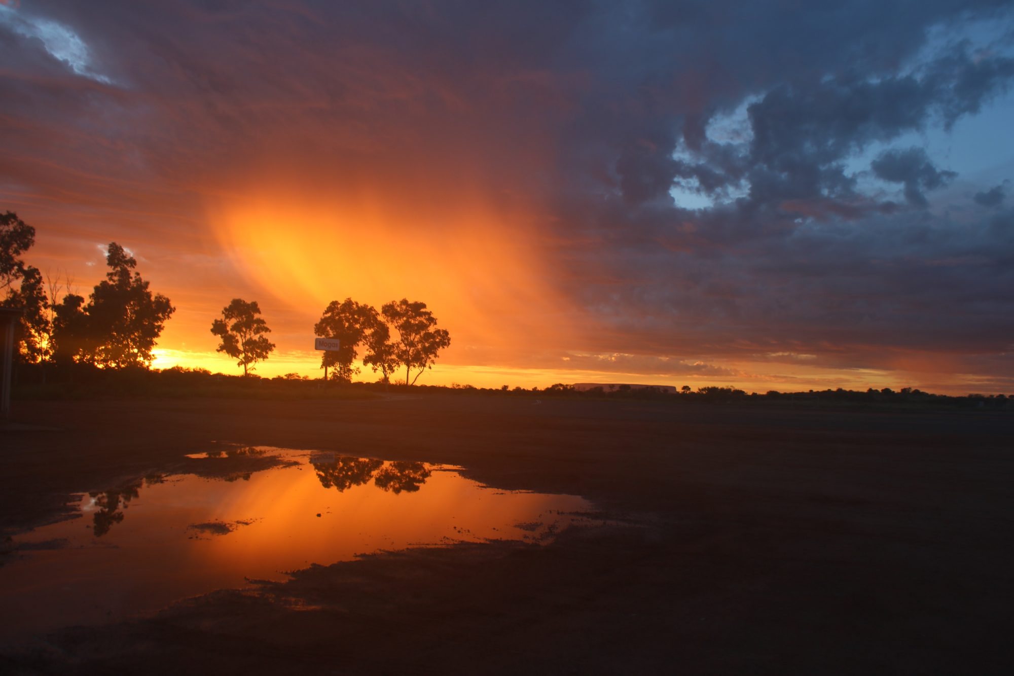 A fiery sunset beneath storm clouds, reflected in rainwater on the plain