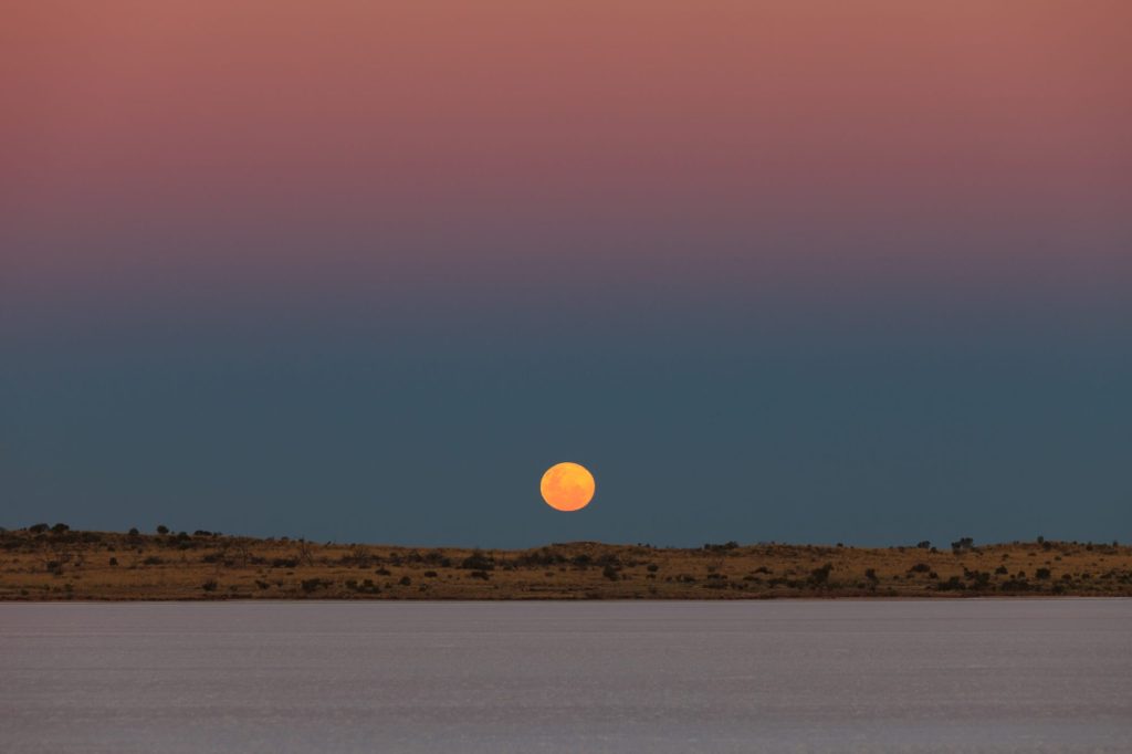 An orange full moon rising over the horizon beyond a pale salt lake at dusk