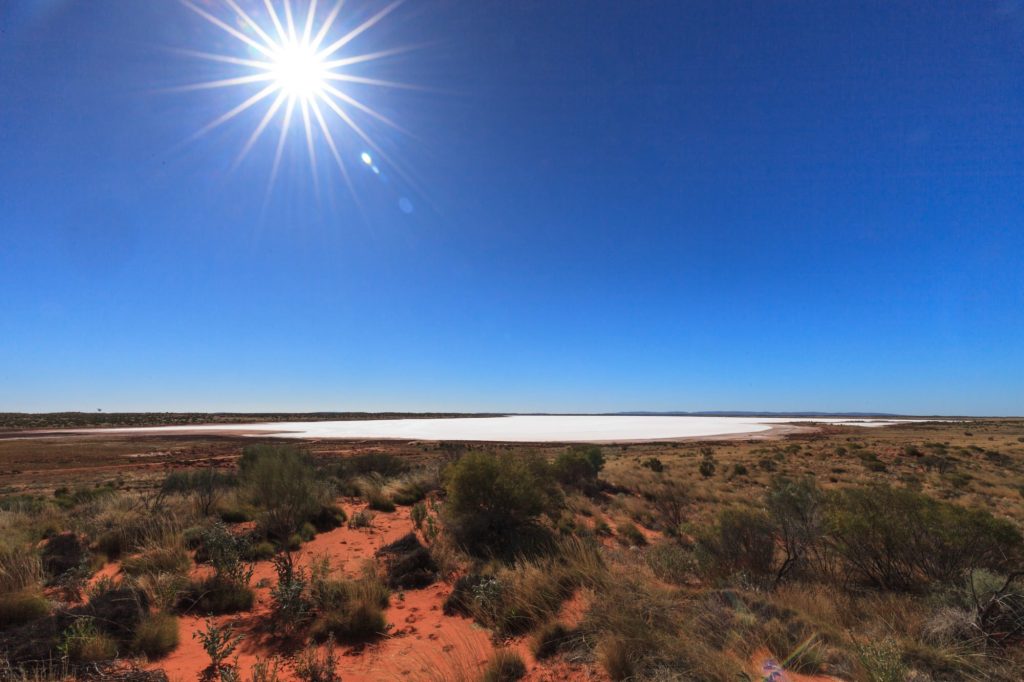 View from a red sand dune over desert scrub to a distant salt lake, with the sun bursting overhead