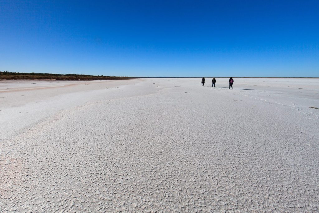 Three small figures walking across a vast white salt lake under a deep blue sky