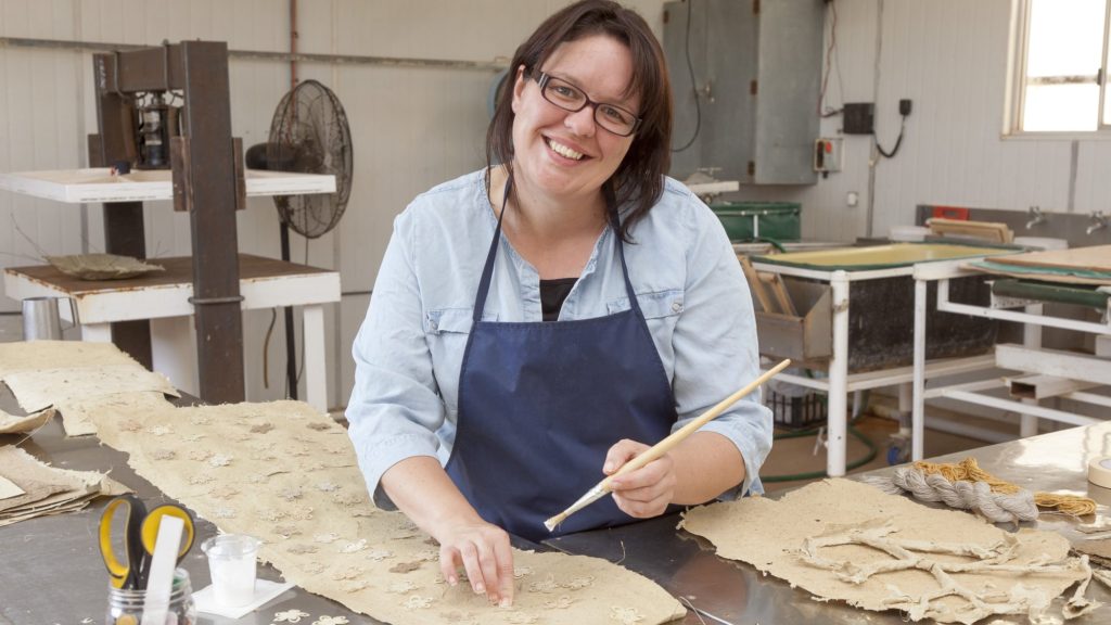 Amee Dennis smiling at her bench in the paper studio among sheets of handmade paper