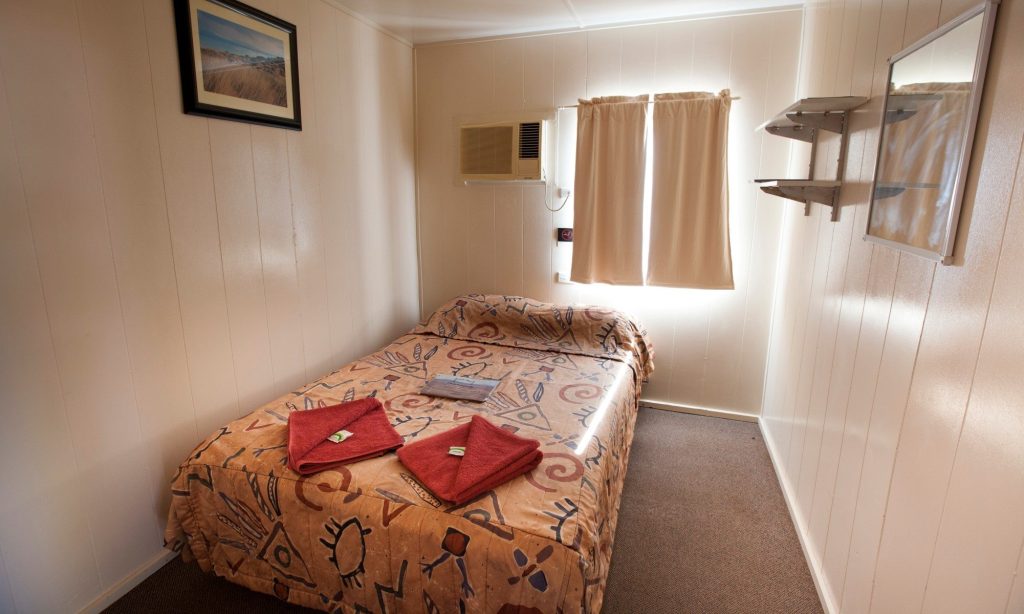 Simple budget room with a made double bed, folded red towels and a wall air conditioner